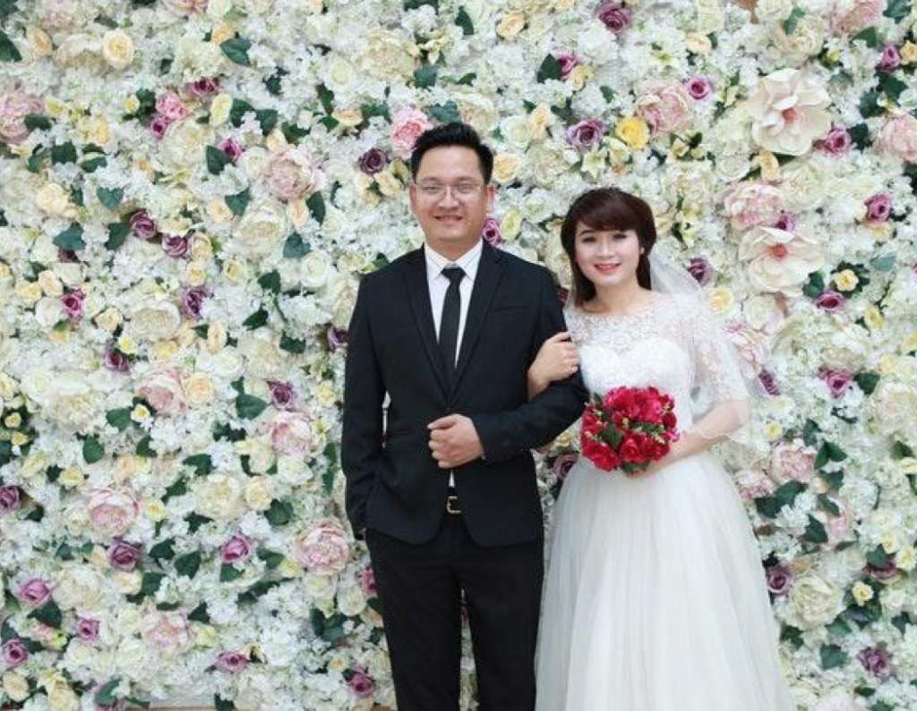 An Asian bride and groom smile in front of a wall of flowers.